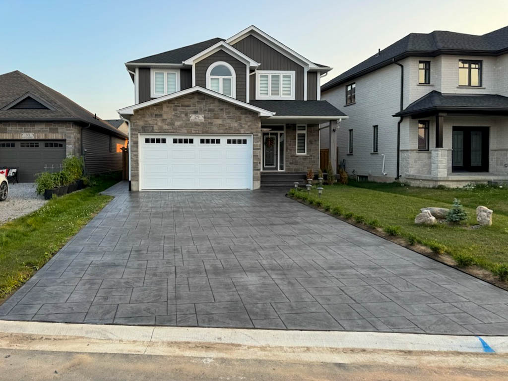 Two-story house with gray siding, stone accents, and a white double garage door, featuring a wide, dark stamped concrete driveway and a small front lawn.
