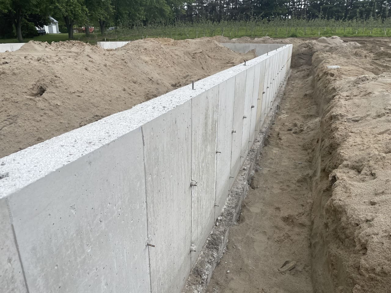 A freshly poured concrete foundation wall with anchor bolts, surrounded by piles of dirt and an adjacent trench at a construction site.