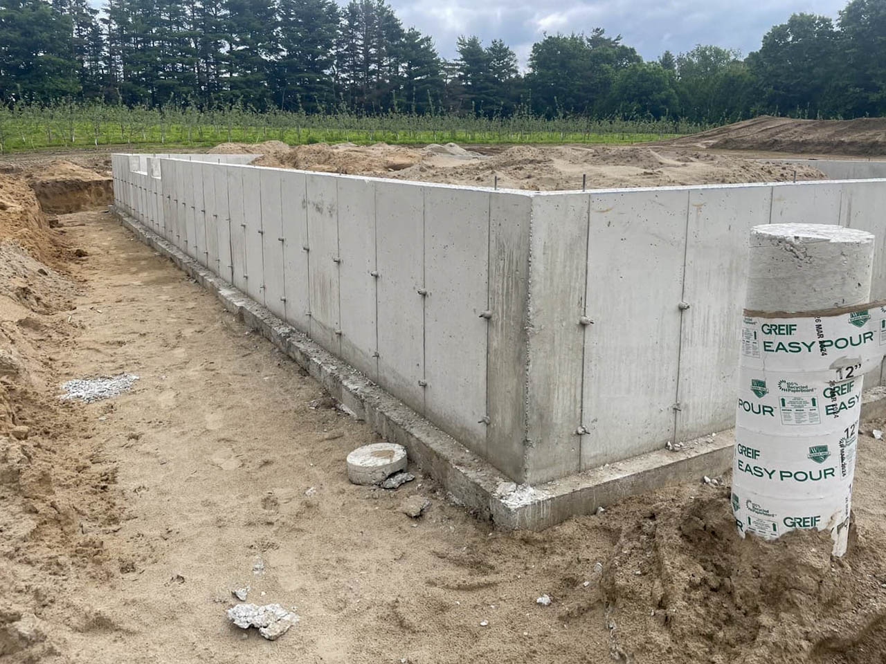 Concrete foundation walls and a circular concrete form are set in a sandy construction site, with trees and greenery visible in the background.