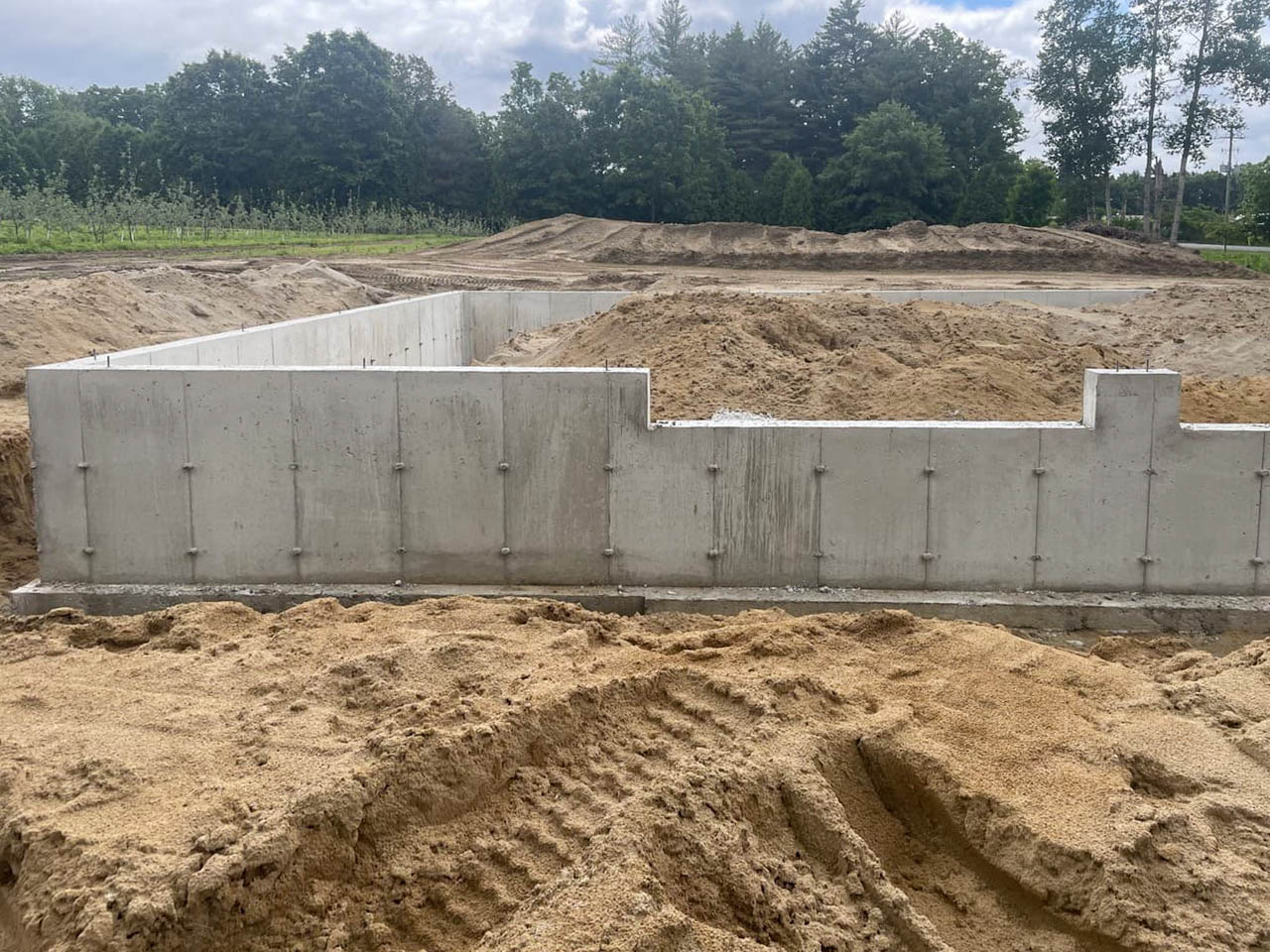 Concrete foundation walls are set in a sandy construction site, with mounds of dirt and trees visible in the background under a partly cloudy sky.