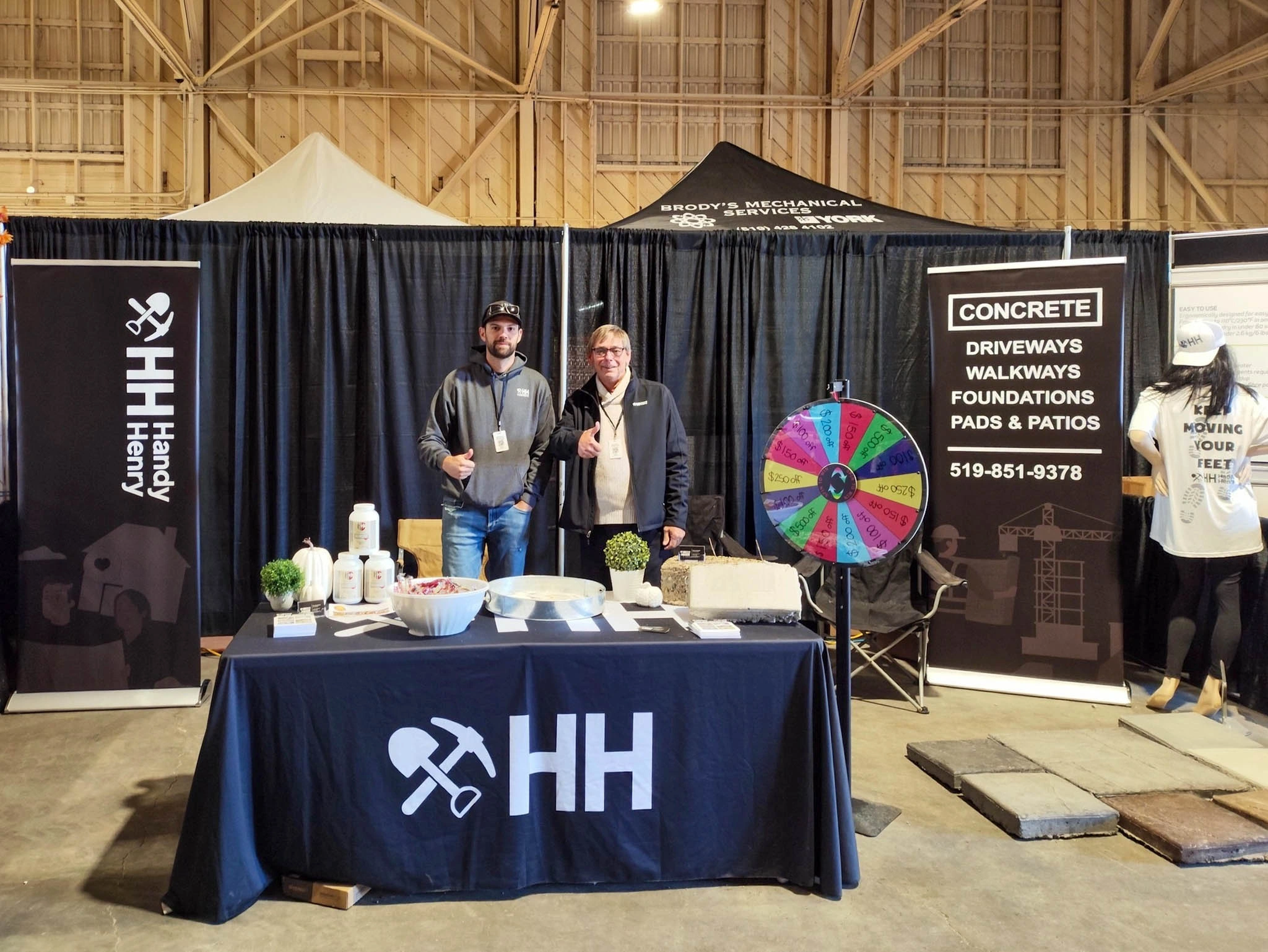 Two men stand behind a Handy Henry booth at an indoor event, with promotional materials, a prize wheel, and concrete service signs displayed.