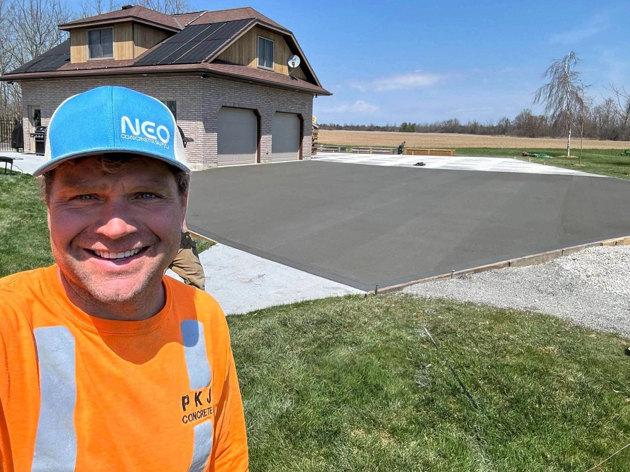 A man in an orange shirt and blue hat stands smiling in front of a freshly poured concrete driveway next to a house with a garage.