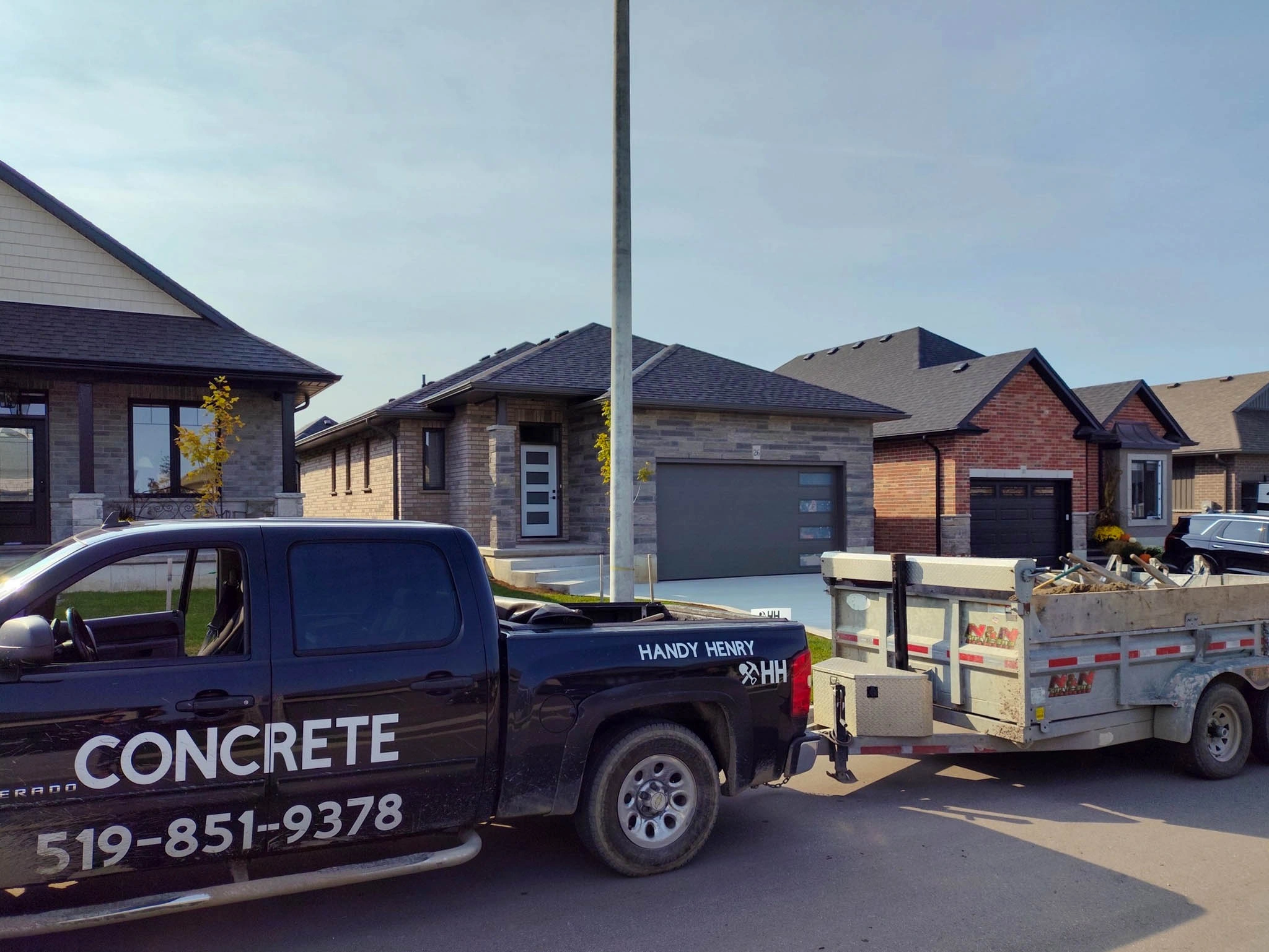 A black pickup truck labeled CONCRETE with a phone number is parked on the street, hitched to a trailer carrying construction debris in front of modern suburban houses.