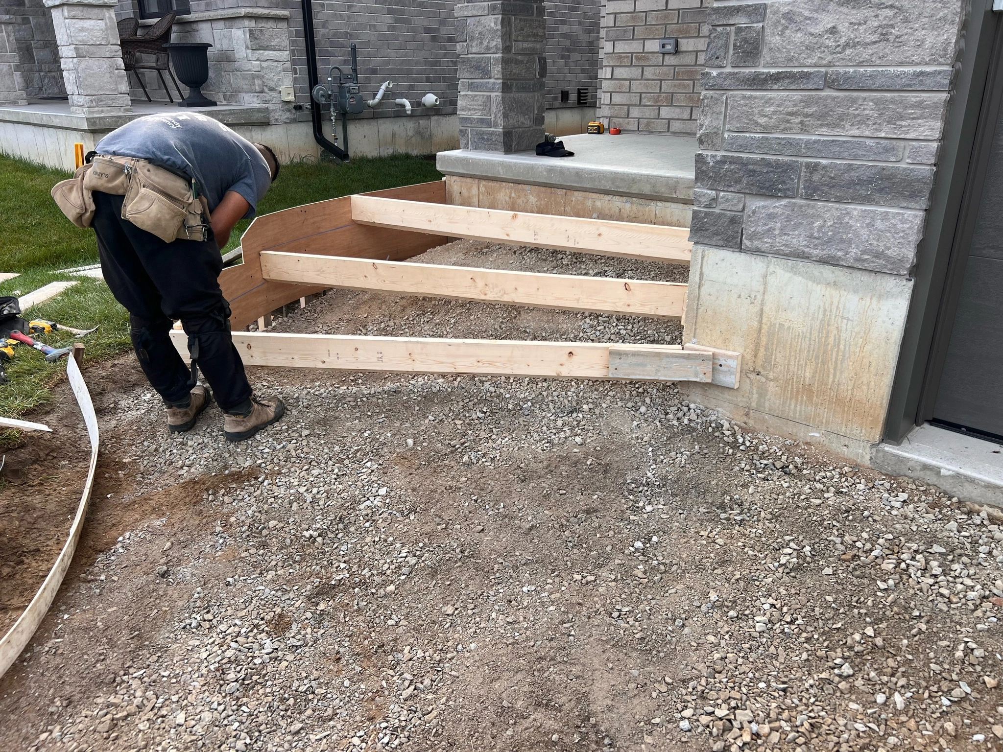A person assembles wooden forms for steps outside a brick house, preparing the area for concrete pouring. Tools and construction materials are visible nearby.