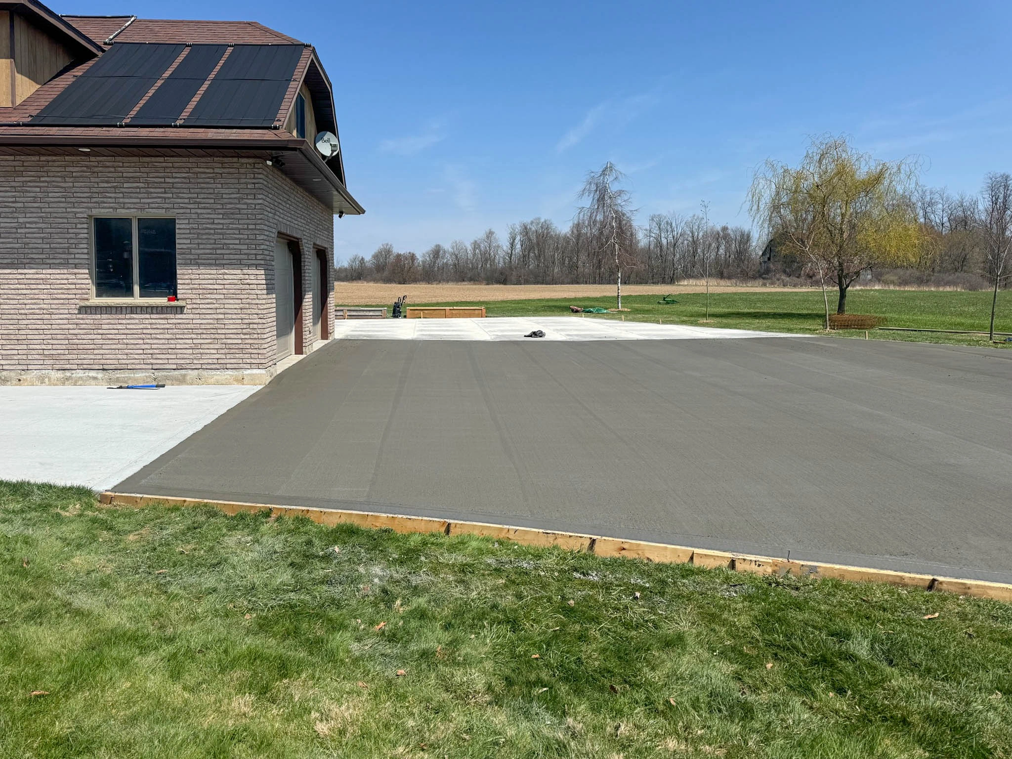 Freshly poured concrete driveway next to a house with solar panels, bordered by grass and trees in the background.