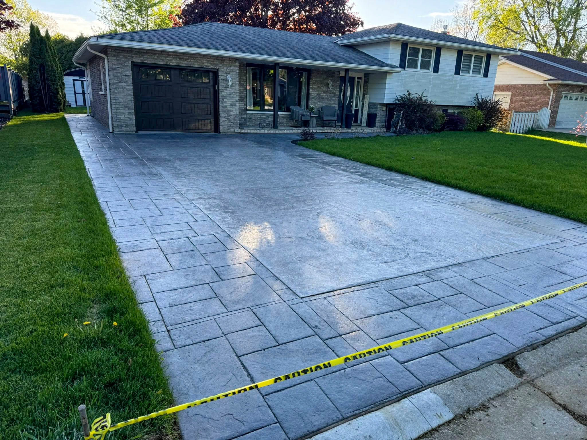 A newly poured and stamped concrete driveway with caution tape blocks access in front of a suburban house with a lawn and porch.