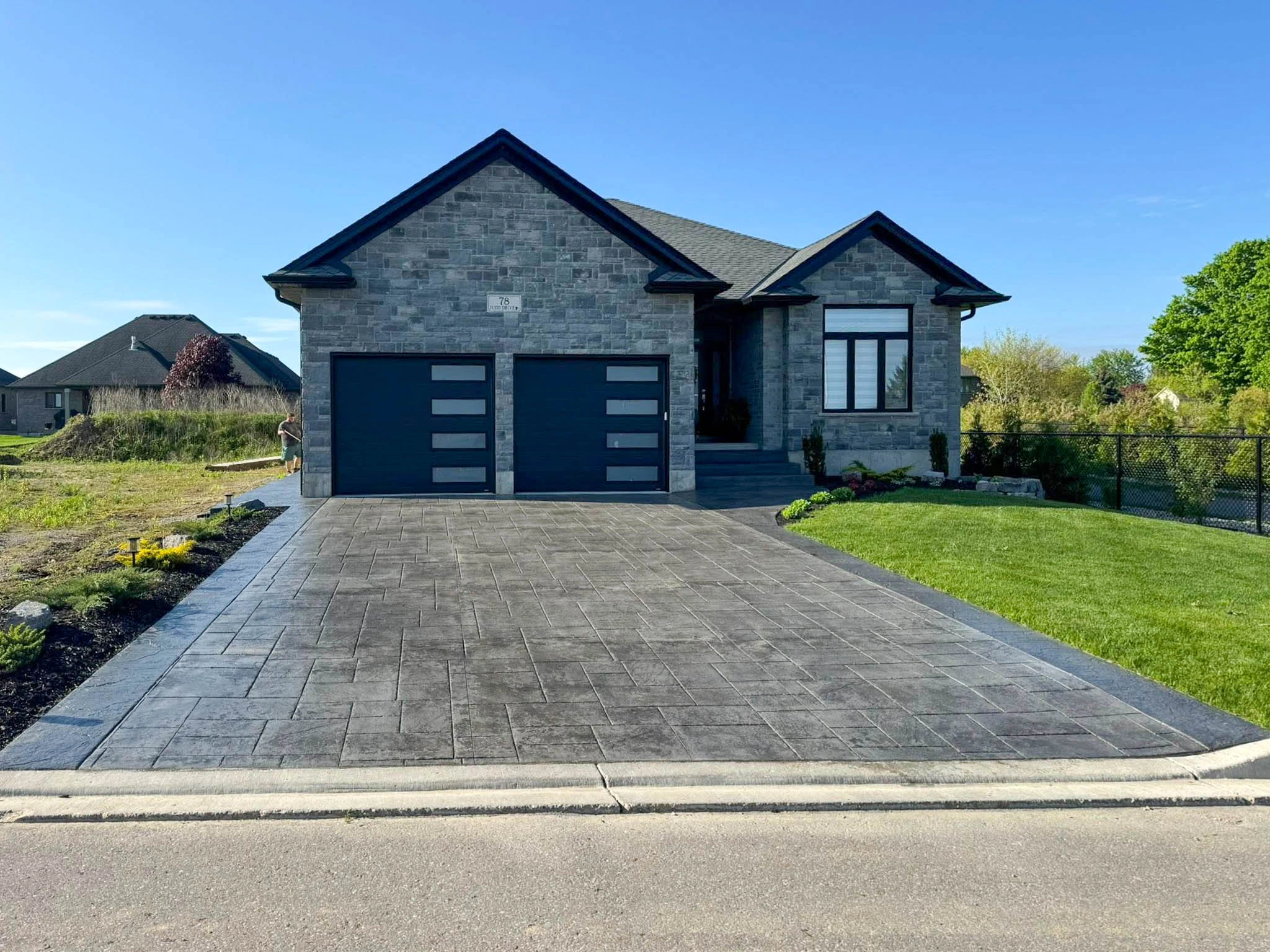 Modern single-story house with a grey brick exterior, two-car garage, and a wide stamped concrete driveway, set in a suburban neighborhood on a clear day.