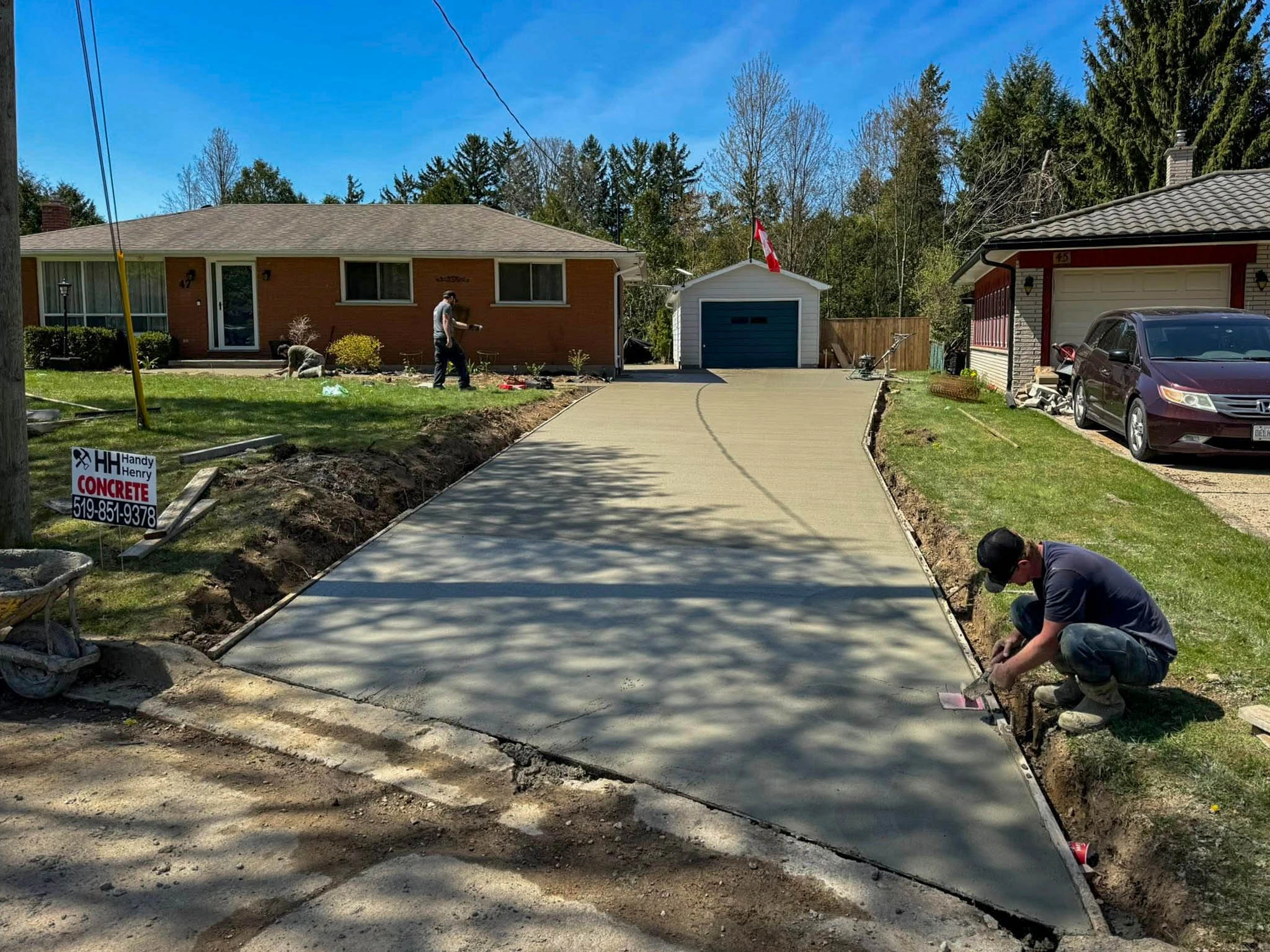 Two workers smooth freshly poured concrete on a residential driveway in front of a brick house with a blue garage.