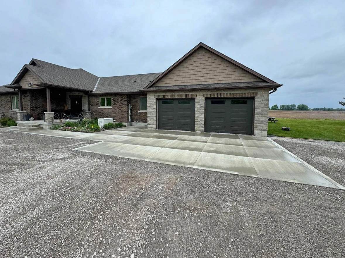 A single-story brick house with a double garage and a wide concrete driveway, surrounded by gravel and lawn, under a cloudy sky.