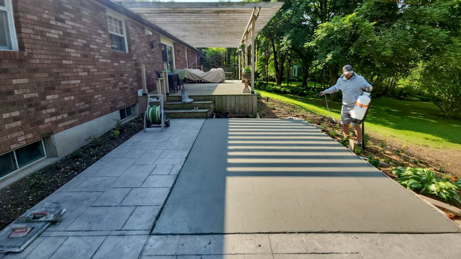 A person applies finishing touches to a freshly poured concrete patio next to a brick house and wooden deck in a backyard.