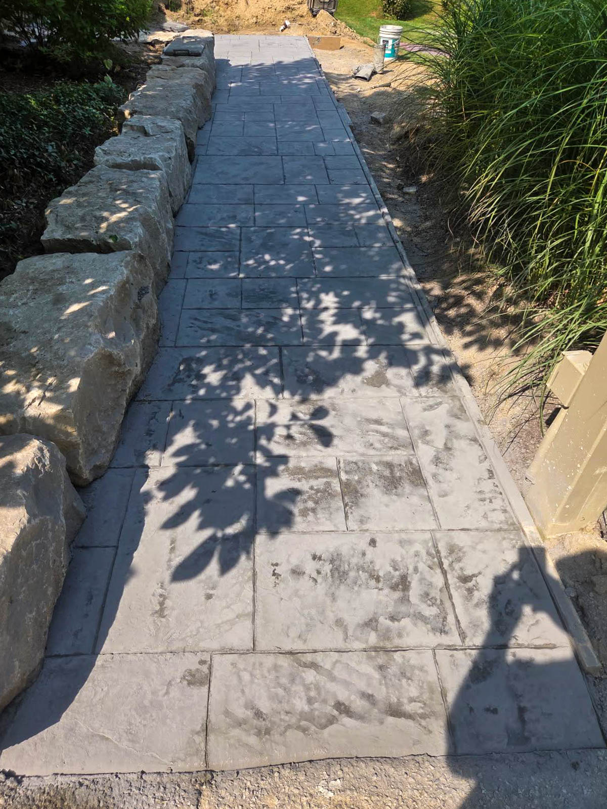 A newly installed stamped concrete walkway bordered by large rocks on one side and tall grass on the other, with tree shadows cast across the path.