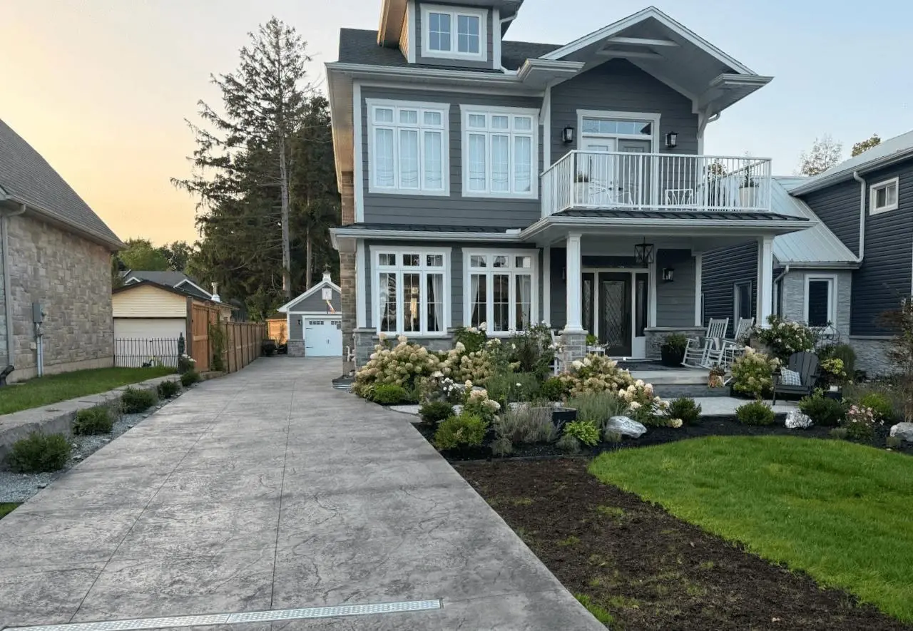 Two-story gray house with white trim, a balcony, large windows, landscaped front yard, and a long concrete driveway leading to a standalone garage.