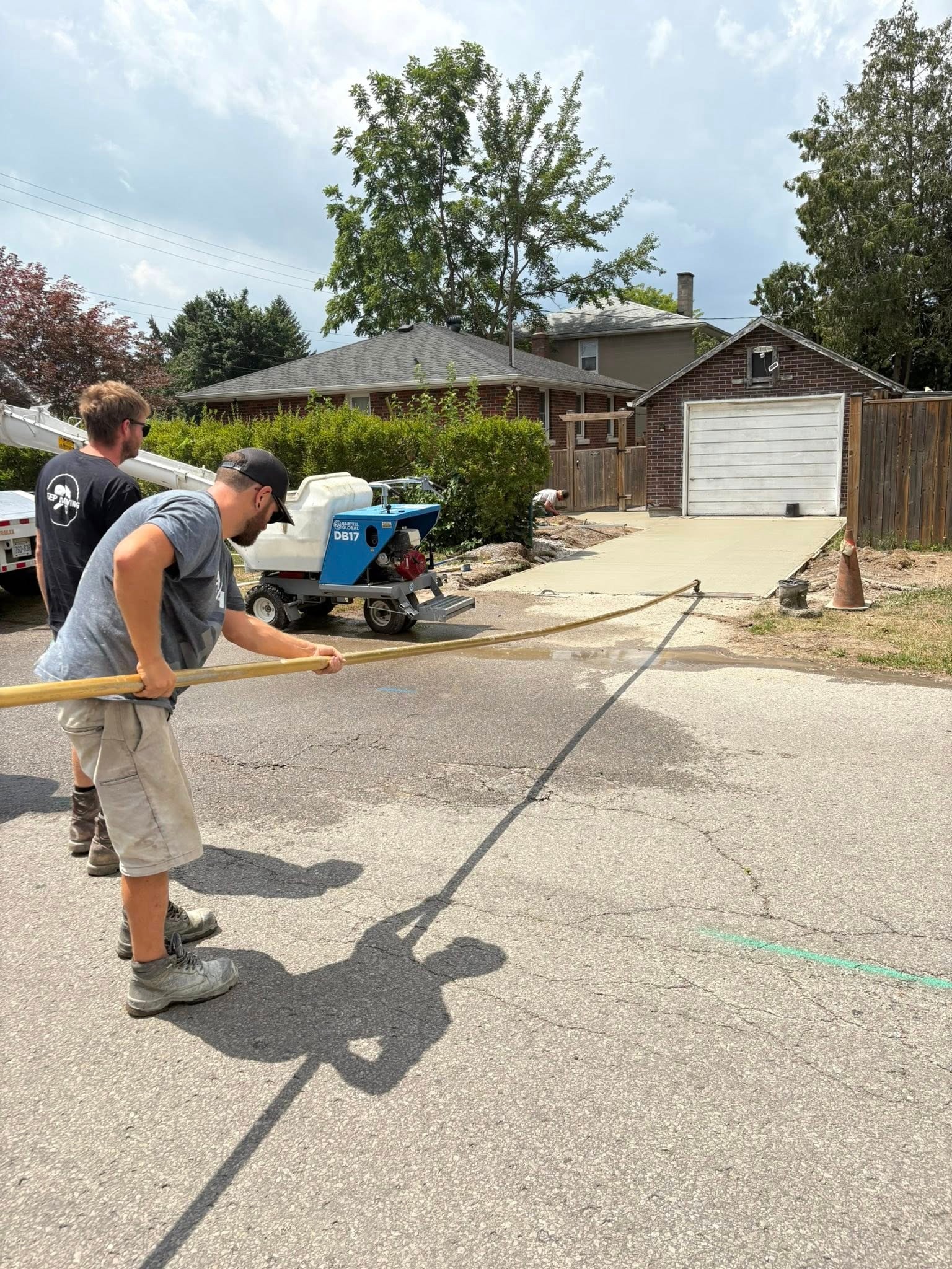 Two workers use a long tool to spray or clean a crack in the pavement on a residential street near a driveway and a parked construction machine.