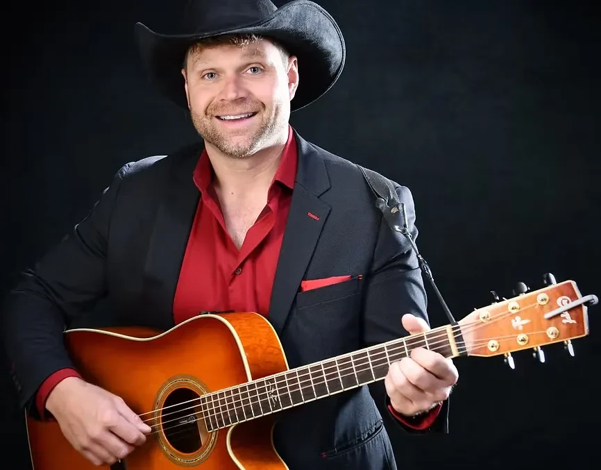 A man wearing a black cowboy hat and suit with a red shirt plays an acoustic guitar and smiles against a black background.