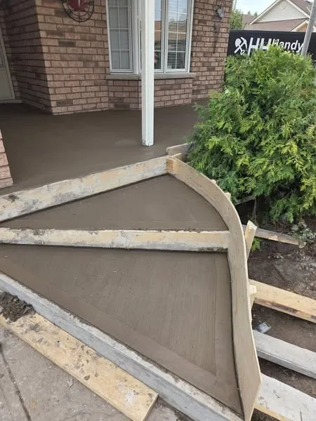 Freshly poured concrete sidewalk with wooden forms in place, leading to the steps of a brick house with a shrub nearby.