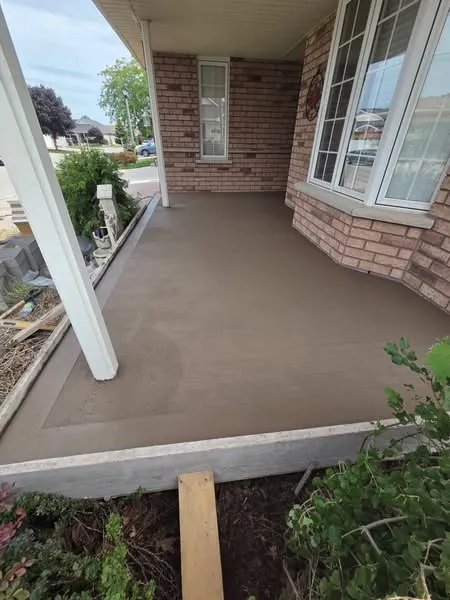 Freshly poured and smoothed concrete porch in front of a brick house, with a garden bed and street visible in the background.