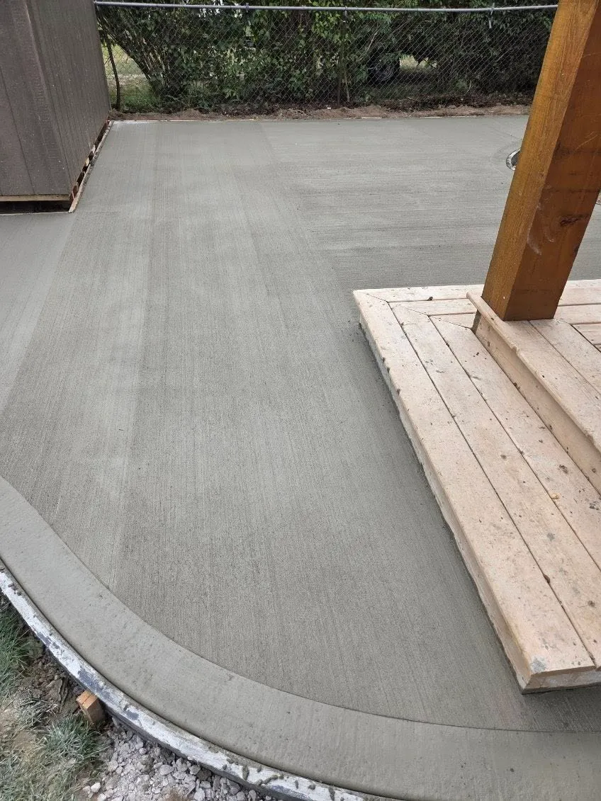 A freshly poured and smoothed concrete slab surrounds a wooden post and structure, with a curved edge near the foreground and a shed along the left side.