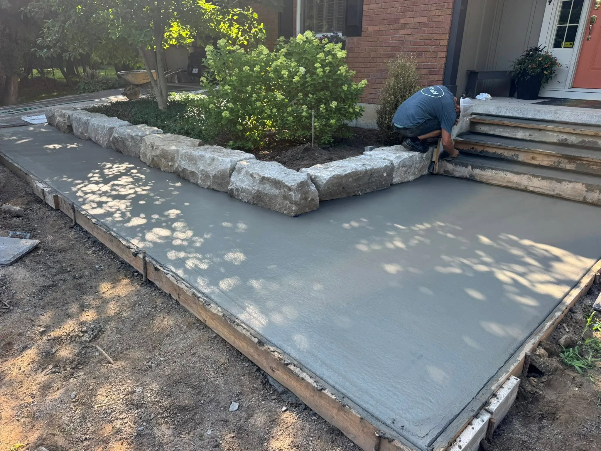 A person smooths freshly poured concrete on a walkway next to stone edging and garden beds in front of a house.